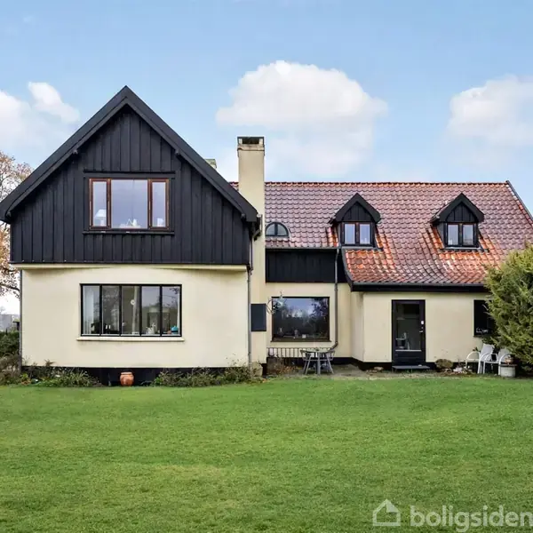 Et to-etagers hus står stille i en grøn have med en blå himmel bagved. Huset har sort træbeklædning, gule vægge og et rødt tegltag.