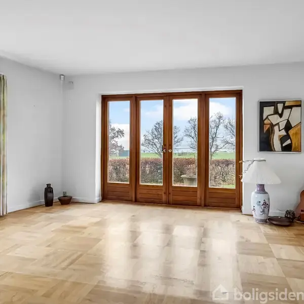 Et lyst rum med trægulv og store vinduer med udsigt til træer. En guitar, en lampe og vaser står ved siden af et abstrakt maleri.