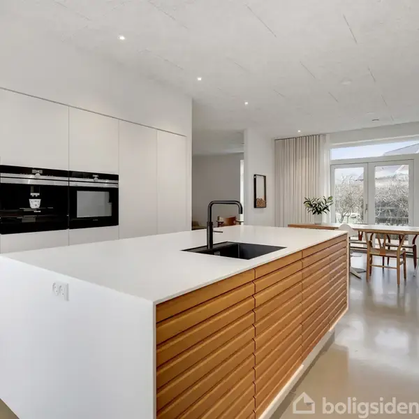 Et moderne køkken med en lang køkkenø har en vask og indbygget ovn. Det åbne rum fører til en spisestue med spisebord og stole nær store vinduer.