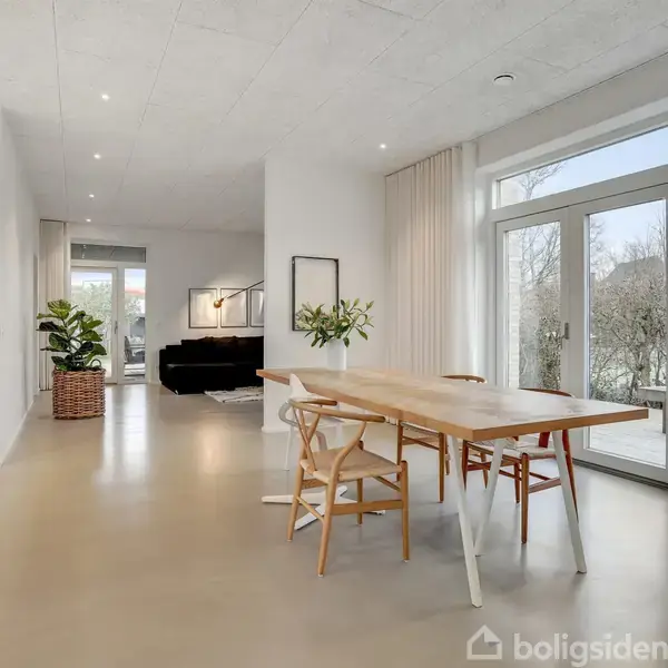 Et spisebord står i et moderne, lyst rum med store vinduer, der giver udsigt til en have. En plante og en sofa ses i baggrunden.