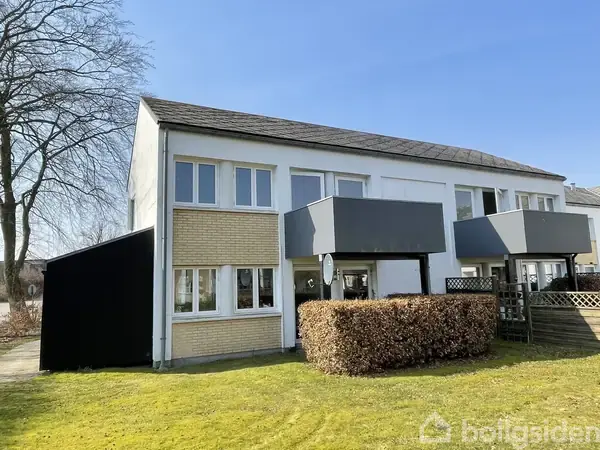 Tofags rækkehus med grå altaner, omringet af en velplejet græsplæne og tætklippede buske. Klare blå himmel og træer i baggrunden.