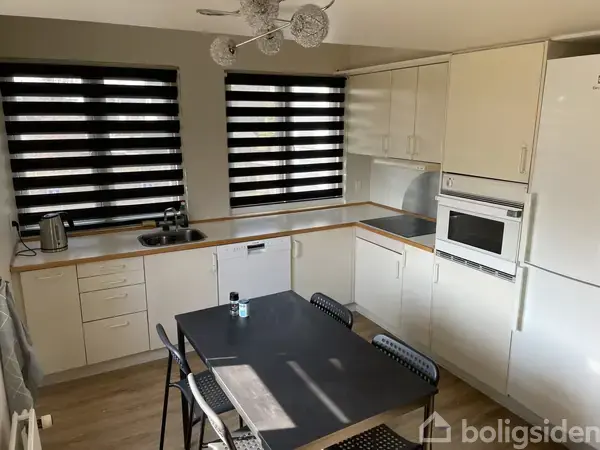 Et lyst køkken med hvide skabe, hvor et sort bord med fire stole står centralt. Der er en mikrobølgeovn indbygget over en ovn, og vinduerne har sorte persienner.