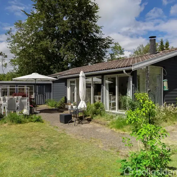Et mørkt træhus med store vinduer står i en frodig have med havemøbler og parasoller. Omkranset af træer og buske under en delvis skyet himmel.