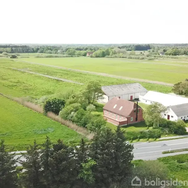 En rød bygning med tilstødende hvide lader står i en grøn landbrugsmark ved en vej. Omkringliggende træer og marker strækker sig mod horisonten.