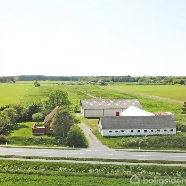 En gård set fra oven består af et rødt hus og to hvide staldbygninger. Bygningerne er omgivet af grønne marker og ligger ved en vej.
