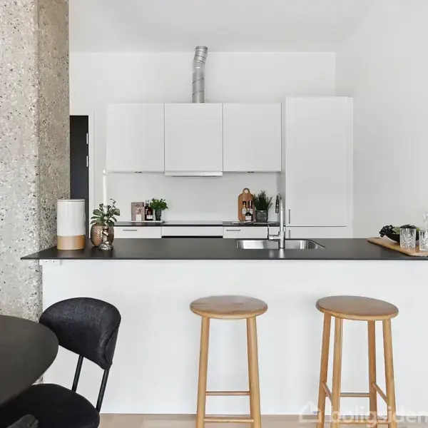 Et moderne køkken med hvidt interiør og sort bordplade. To høje stole står ved køkkenøen. Der er planter, en vandhane, og en bakke med glas. En betonvæg ses til venstre.