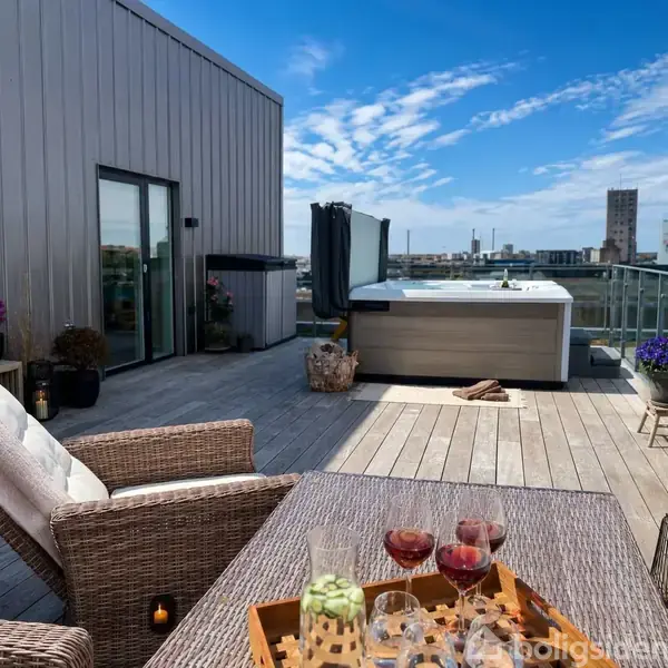 Terrasse med spisebord, hvorpå der står drikkevarer og snacks. Luksuriøst boblebad i baggrunden. Omgivet af byudsigt og dekorative planter under en klar, blå himmel.