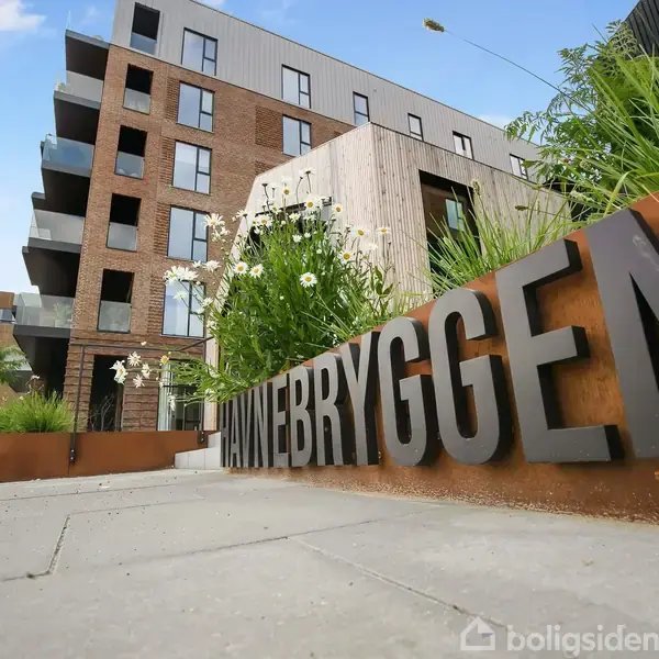 Moderne lejlighedsbygning med flere etager og altaner. Foran bygningen er en stor plantekasse med grøn beplantning og teksten "HAVNEBRYGGEN" i fremtrædende bogstaver langs kanten.