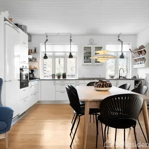 Et lyst køkken med træbord og sorte stole står midt i rummet. Ved bordet er der en moderne pendellampe. Køkkenet har hvide skabe, komfur, ovn og store vinduer.