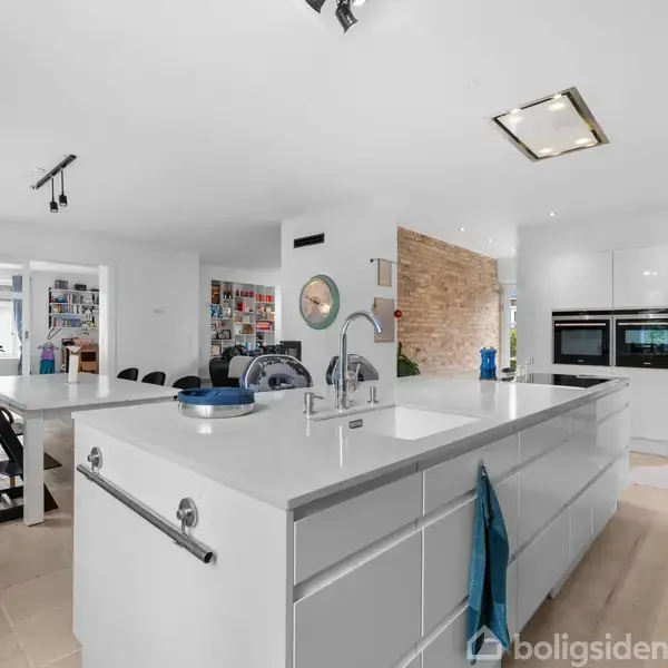 Et moderne køkken med hvidt køkkenø, dobbelt vask og indbyggede ovne. Omgivet af spiseområde med glasvitrine og åbne trapper. Lyse, rummelige omgivelser med minimalistisk indretning.