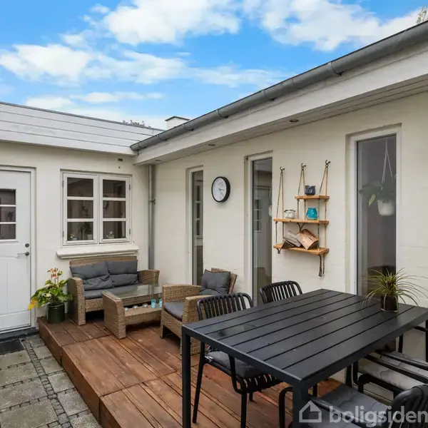Terrassemøbler arrangeret omkring et sort bord på en terrasse af træ. Rundt om er der hvide husvægge med et dør- og vinduesparti; dekoreret med planter og en væghylde.