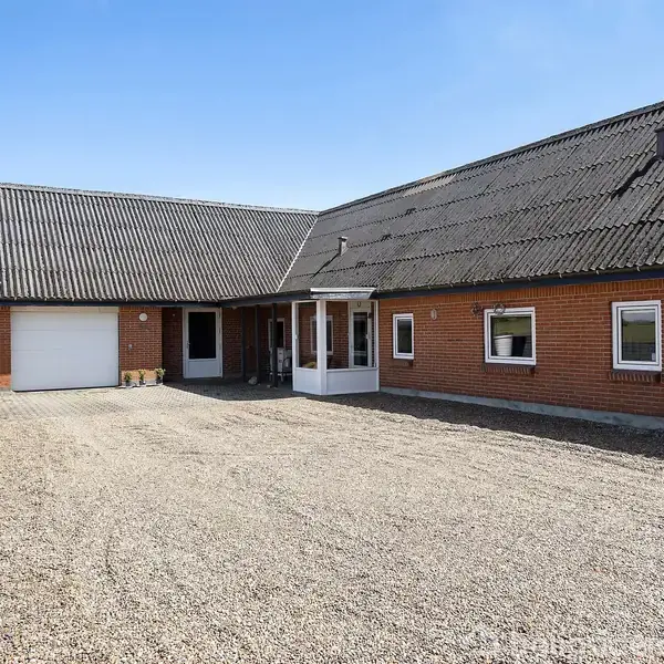 En murstensbygning med sort tag står i en grusbelagt gårdsplads. Bygningen har flere vinduer og en garageport. Der er plantekrukker langs en væg under en blå himmel.