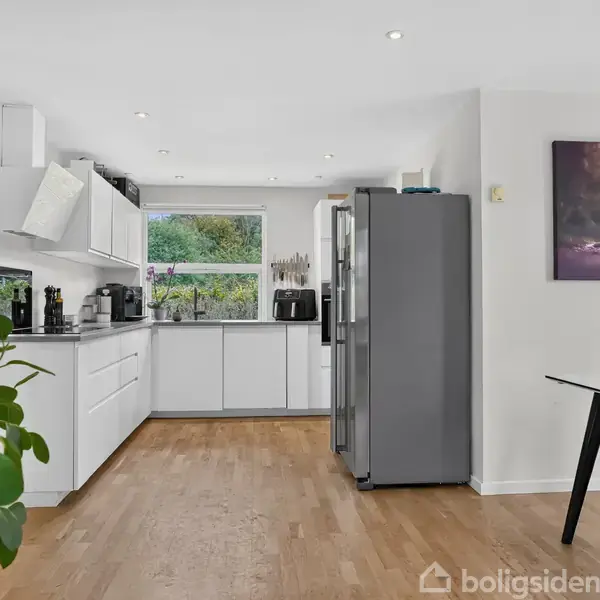 Et moderne køkken med hvide skabe og sorte apparater står stille. Ved siden af er et spisebord med plexiglasstole. En stor plante står i venstre side, mens et stort vindue bagved giver udsigt til en have. En kunstplakat hænger på væggen.
