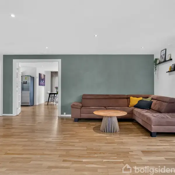 En brun, L-formet sofa med puder står mod en grøn væg i en stue med trægulve. Et rundt træbord står foran sofaen. Hylder med billeder er på væggen.