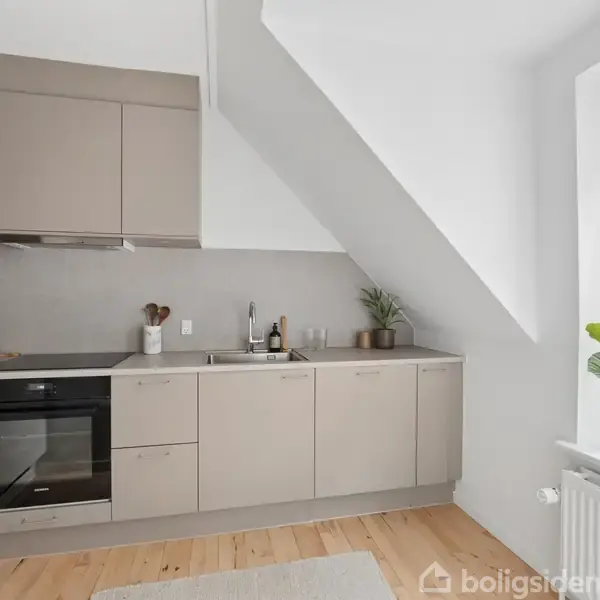 Et moderne køkken med beige skabe udfører opbevaringsfunktioner. Et komfur og en ovn er integreret. Rummet har et stort vindue med udsigt til hustage. Planter pynter bordpladen.