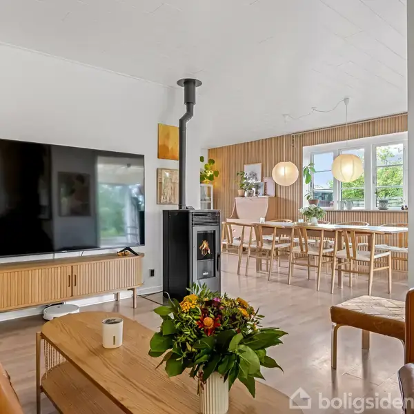 Et moderne stueområde har en brun sofa, et sofabord med en blomsterbuket, en stor TV på et skab, en pejs, og i baggrunden et spisebord med stole ved et vindue.