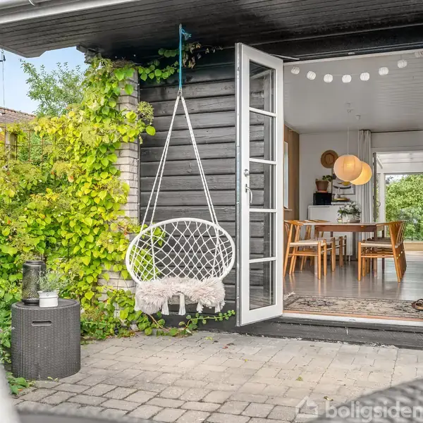 Gyngestol hænger udenfor en åben dobbeltglasdør til en stue. Indenfor er der spisebord og lamper. Omkring er grønne planter og en rummelig terrasse med fliser.