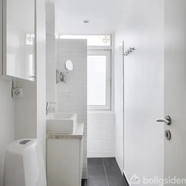 En lang, hvid lejlighedsbygning står på en velplejet grøn plæne. Bygningen har flere etager og balkoner. Himlen er blå med enkelte skyer, og området er omgivet af træer.