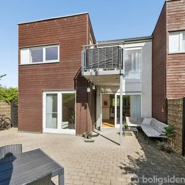 To-etagers moderne hus med træfacade har store vinduer og balkon. Udenfor er der en møbleret fliseterrasse omgivet af en bambushegn og frodige buske.