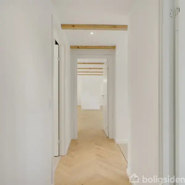En lang, hvid gang strækker sig fremad, flankeret af døre på begge sider. Lyse trægulve danner et mønster. Træplader på loftet bryder den ellers glatte overflade.