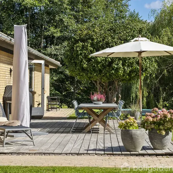 En terrasse med træmøbler og parasol står foran et gult murstenshus. Omkring er der grønne buske og træer. En stol og blomsterkrukker tilføjer hyggelig atmosfære.