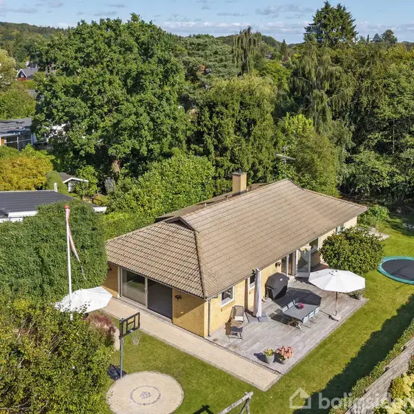 Huset er en gul murstensvilla med rødt tegltag. Det står i en grøn have med en terrasse, parasol, trampolin og omgivende træer. Baggrunden er fyldt med frodig skov.