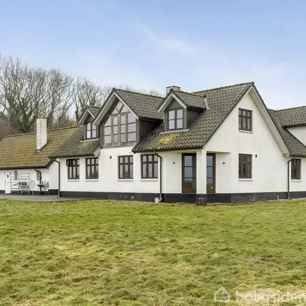 Hvidt hus med gråt tag står på en åben, grøn græsplæne. Omkring huset er der træer og en klar blå himmel.