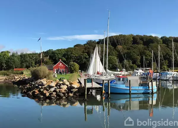 Både ligger til kaj i en lille havn, stille vand. Omkring er der grønne træer og en rød træbygning med en grusvej. Skilt foran broen.