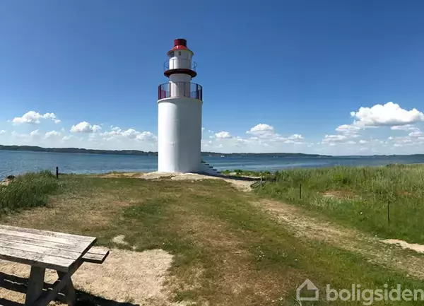Fyrtårn står ved kysten under en blå himmel med få skyer. Græs og en træbænk omgiver området, og vand strækker sig ud i baggrunden.