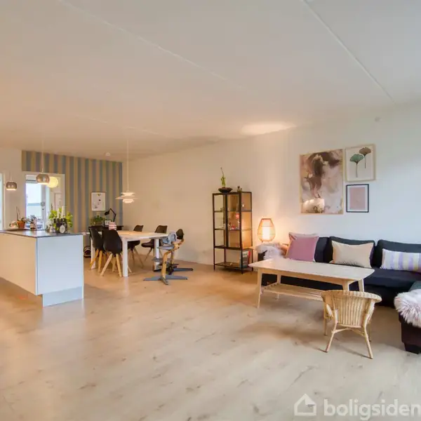 En stue med en blød, mørk sofa fyldt med puder står i et åbent rum. Ved siden af sofaen er et sofabord og en kurvestol. Køkkenet ses i baggrunden med spisebord.