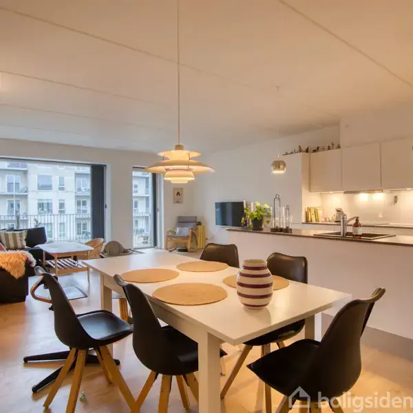Spisebord med stole står centralt; lyskroner hænger over det. Køkken med hvide skabselementer til højre. Stue med sofa og hylde til venstre; store vinduer i baggrunden.