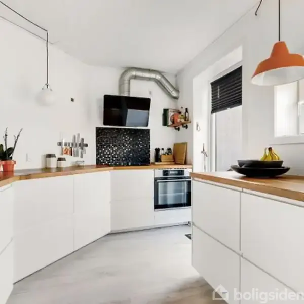 Et moderne køkken med hvide skabe og træbordplader. En kogeplade og emhætte er midt på bagvæggen. Rummet er lyst med planter og hængende lamper.