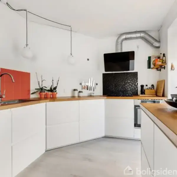 Et moderne køkken med hvide skabe og træbordplader. Der er en ovn og emhætte samt planter og dekorationer på bordet. Køleskabet har magneter, og rummet er lyst.