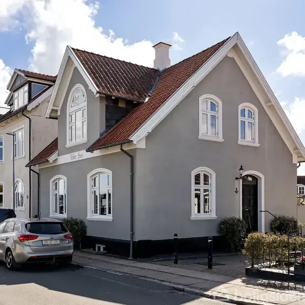 Et gråt hus med rødt tag står på en byvej, omkranset af biler og en blå himmel med hvide skyer.