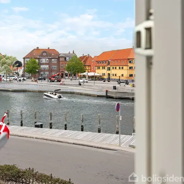 Udsigt fra vindue til en båd sejler på en kanal i bymiljø med farverige bygninger og træer i baggrunden. En dansk flag er synligt i forgrunden.