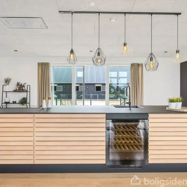 Køkken med stor ø i forgrunden med trælister og integreret vinkøler. Over øen hænger moderne lamper. Bagved ses store vinduer med udsigt til huse. Ingen tekst er til stede.