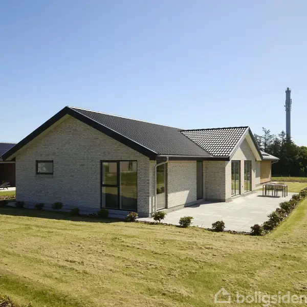 Et moderne enfamiliehus med mørkt tag og lyse murstensvægge står stille i en stor, grøn græsplæne. Der er en terrasse på siden og træer i baggrunden.