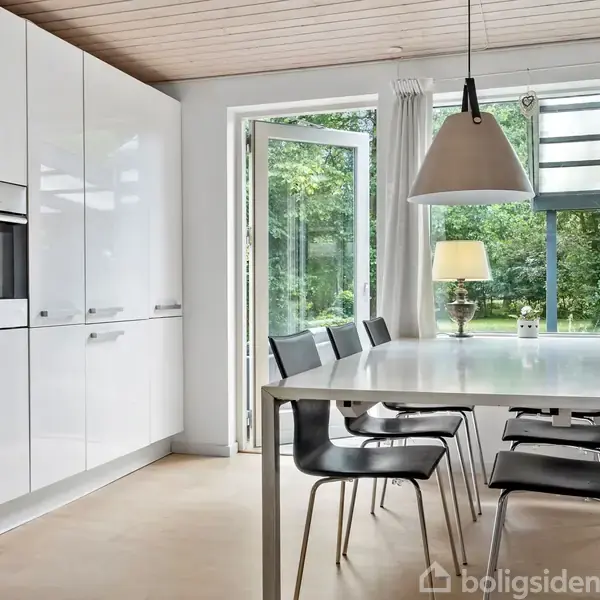 Køkken-alrum med hvidt spisebord omgivet af syv sorte stole. Bordlampe og glasfyldte døre fører ud til en grøn have. Højskabe til venstre med indbygget ovn.