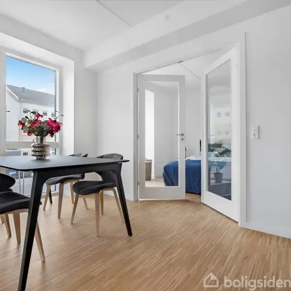 Spisebord med sorte stole står i et lyst rum med trægulv. En vase med røde blomster pryder bordet. Bagved findes dobbeltdøre, der fører til et soveværelse.