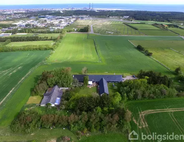 Gårdkompleks omgivet af grønne marker; bygningerne er placeret i et L-formet layout. Landskabet strækker sig mod en by i det fjerne, under en klar blå himmel.