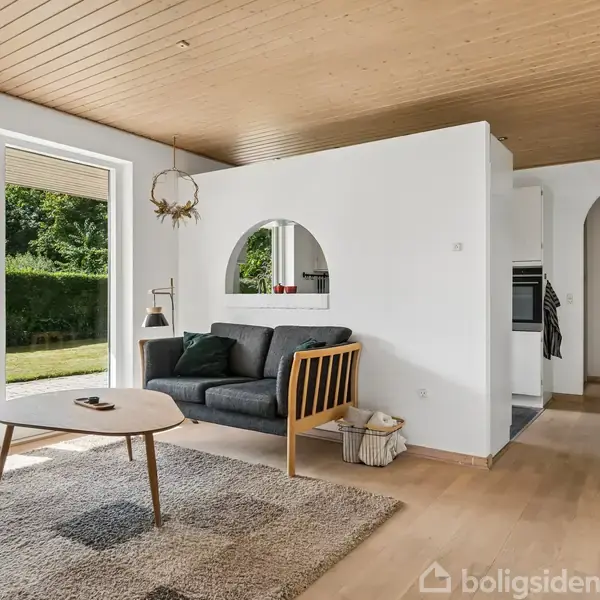 Stue med sofa ved væg og rundt bord på gulvtæppe, store vinduer til venstre giver udsigt til have. Tæppe og udsmykning skaber en hyggelig atmosfære i rummet.