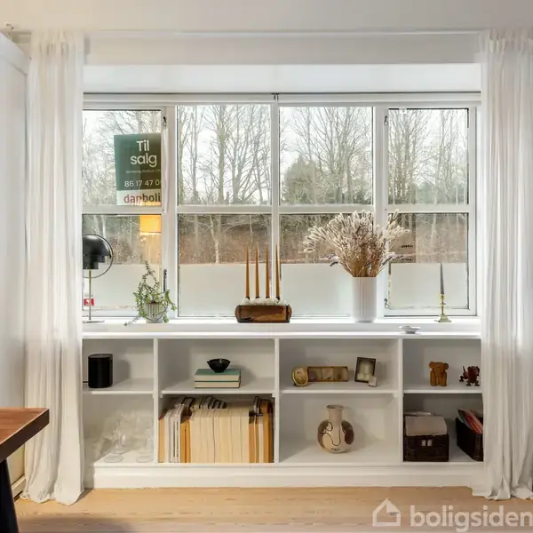 Reol med bøger og dekoration står under et stort vindue. Skilt med teksten "Til salg" hænger på vinduet. Rundt omkring er der et bord, stol og tv.
