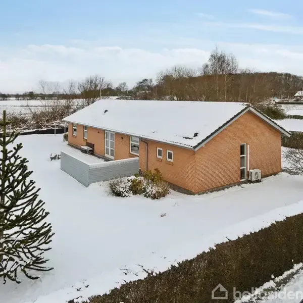 Et murstenshus er dækket af sne og omgivet af en vinterlandskab med hække og et grantræ i forhaven. En parkeret bil ses i hjørnet.