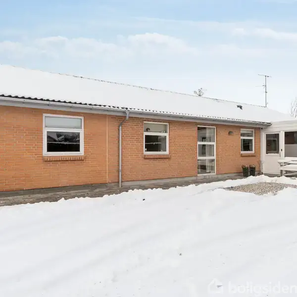 En enkelt etage, rød murstenshus med store vinduer står stille. Sne dækker grunden rundt om huset, og der er en tilhørende terrasse med hvidt rækværk.