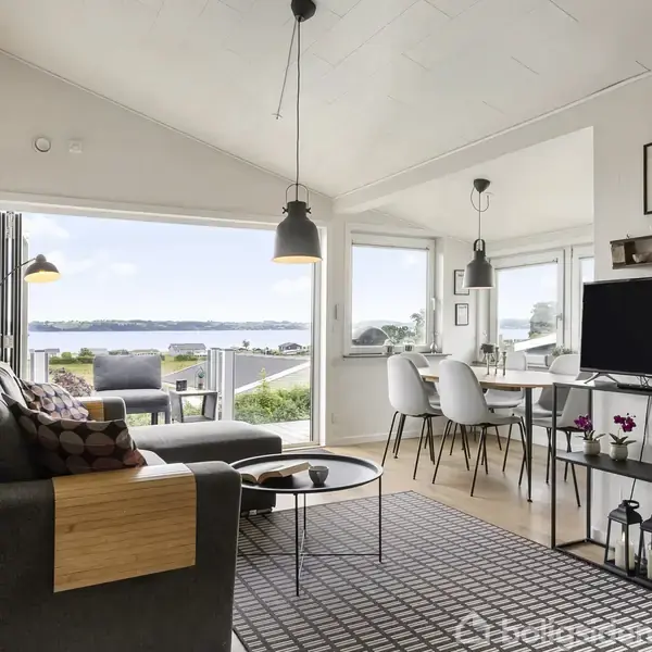 Stue indeholder en grå sofa med puder, som vender mod et sofabord. Ved siden af er en spiseplads med fire stole. Store vinduer viser udsigt over en terrasse og vandet udenfor.