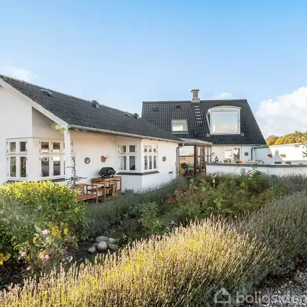 Hvid murstenshus med sort tag har en terrasse med havemøbler. Det ligger i en frodig have med buske og lavendel, omgivet af klar blå himmel og solskinsvejr.