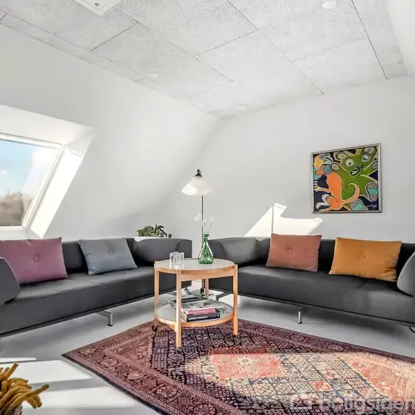 Et sort sofaarrangement står i hjørnet af en lys stue med et skråtag. Et lille rundt bord med bøger og en lampe er i midten, omgivet af farverige puder og et tæppe. En abstrakt maleri hænger på væggen.