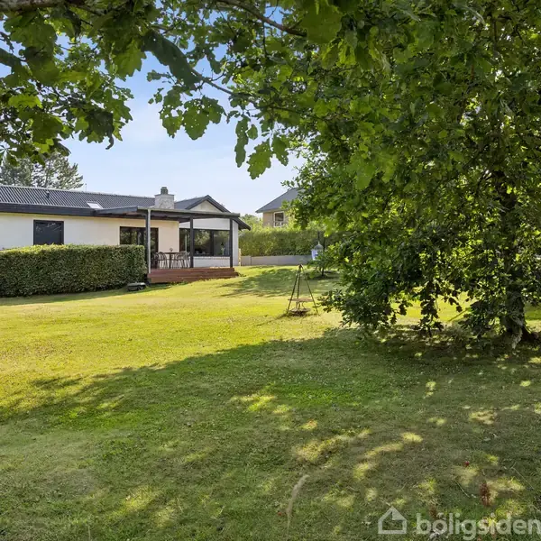 Et hvidt hus med terrasse set fra en grøn have omgivet af buske og træer, der skaber en rolig og indbydende atmosfære.