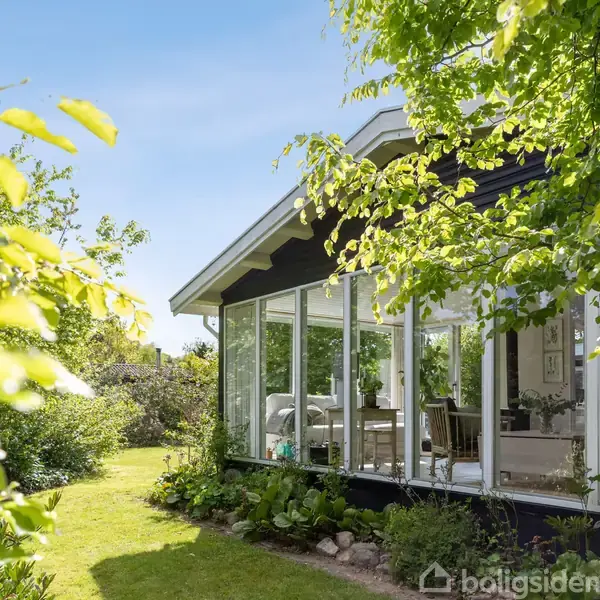 Et hus med store vinduer viser en lys stue. Sollys skinner gennem hængende grene, omgivet af en grøn have.