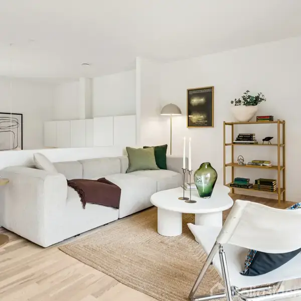 Stue med moderne indretning; hvid sofa med grønne puder ved siden af rundt sofabord. Træhylde med dekorationer, gulvlampe, lys trægulv. Bordlampe og tekande på træsidebord. Ingen tekst.
